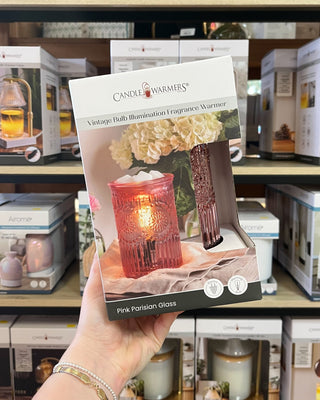 A hand holds a package of Candle Warmers 'Pink Parisian Glass' fragrance warmer in a retail store setting with other