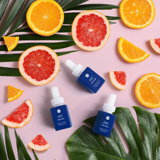 Blue skincare bottles with droppers surrounded by sliced oranges and grapefruits on a pink background