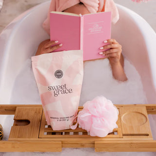 Person in a bathtub with a pink book and 'sweet grace' product, surrounded by bubbles and a bath tray.