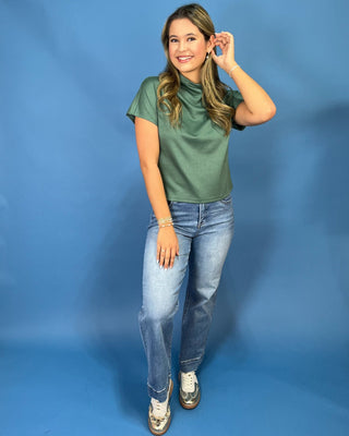 A woman modeling a sage green short-sleeve mock neck top with light-wash blue jeans and white sneakers