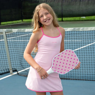 A young girl smiling on a tennis court, wearing a light pink sleeveless tennis dress with bright pink trim and holding a pink-and-white polka dot paddle.